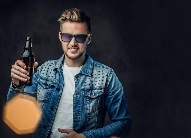 Varón rubio positivo vestido con una chaqueta de mezclilla y gafas de sol sostiene una botella con cerveza artesanal
. - Foto, imagen