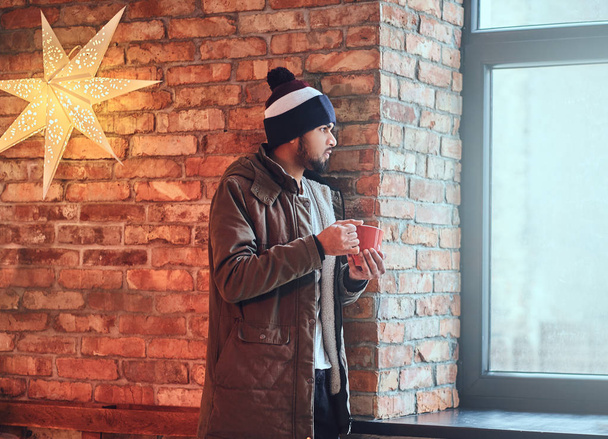 Portret van bebaarde Indiase hipster mannelijke gekleed in een warme jas en een hoed, drinken koffie uit een rode kop in de buurt van de muur van een rode bakstenen. - Foto, afbeelding