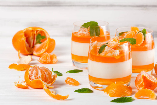 Creamy panna cotta with orange jelly in beautiful glasses, fresh ripe mandarin, on white wooden background. Delicious Italian dessert. Closeup photography. Selective focus. - Foto, imagen