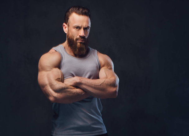 Portrait of brutal bearded bodybuilder with crossed arms dressed in a sleeveless shirt isolated on grey background. - Foto, immagini