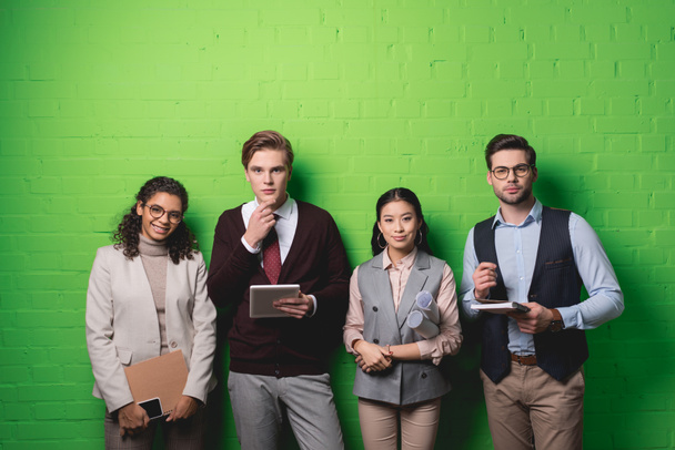 jóvenes empresarios multiétnicos con dispositivos digitales y documentos de pie frente a la pared verde
 - Foto, Imagen