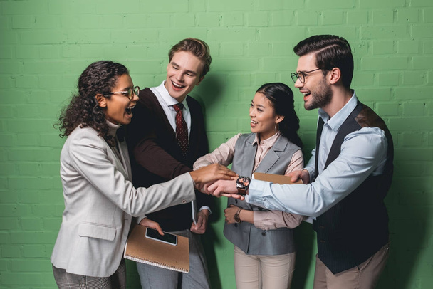 equipo empresarial multiétnico emocionado tomados de la mano frente a la pared verde
 - Foto, Imagen