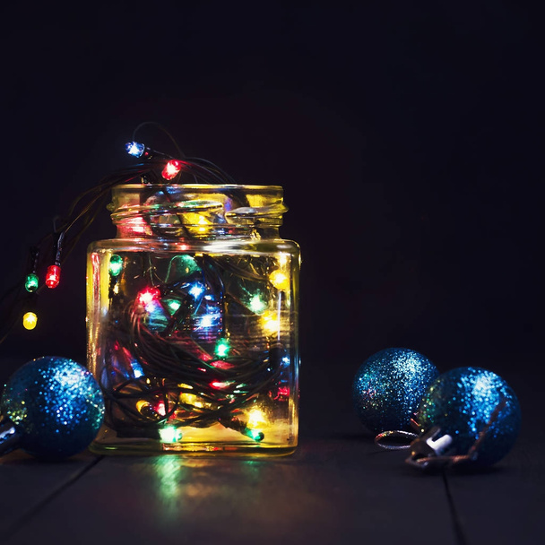 A glowing garland in a glass jar and Christmas decorations on a dark wooden background. New Year, Christmas postcard. Copy space. - Photo, Image