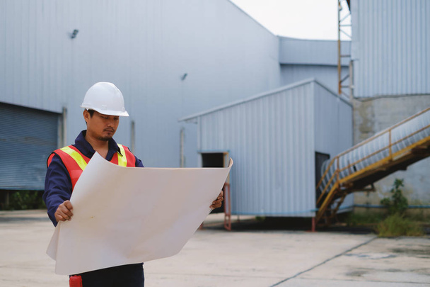 Ingénieur en uniforme de sécurité et casque blanc tenant le plan de projet à l'arrière-plan du chantier de construction en plein air, concept industriel
 - Photo, image