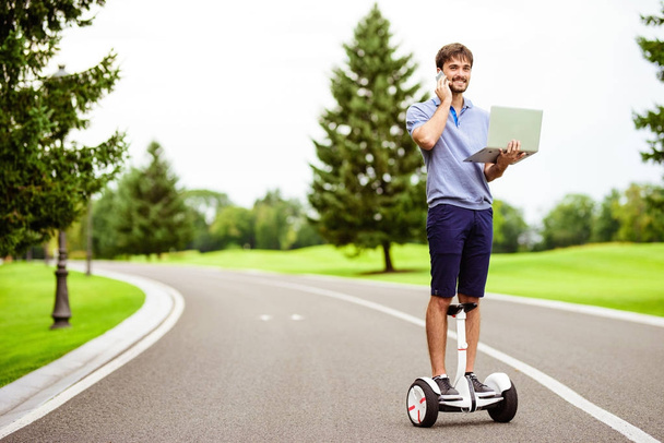 A man is driving through the park on a gyroboard. He holds a laptop in his hands and raschvgovravaet by phone - Фото, изображение