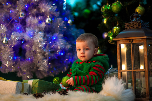 Menino segura bola de Natal na mão e senta-se perto da árvore de Natal
.  - Foto, Imagem