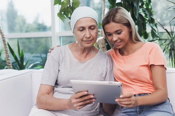Una donna con il cancro è seduta accanto a sua figlia su un divano bianco. Stanno guardando qualcosa su un tablet grigio
. - Foto, immagini