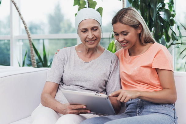 A woman with cancer is sitting next to her daughter on a white sofa. They are looking at something on a gray tablet. - Foto, Imagem