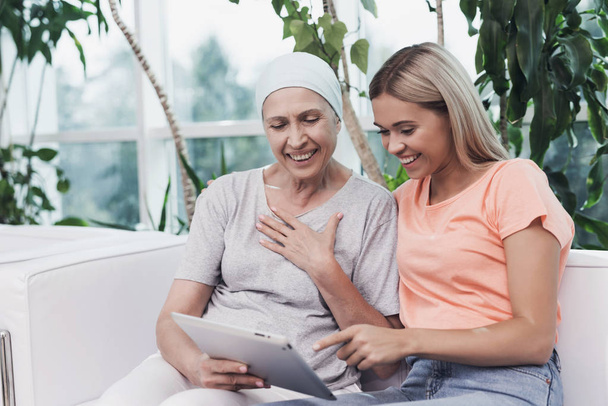 A woman with cancer is sitting next to her daughter on a white sofa. They are looking at something on a gray tablet. - Photo, image