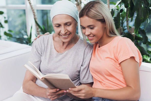A woman with cancer is sitting next to her daughter on a white sofa. They are looking at something in the book. - Photo, Image