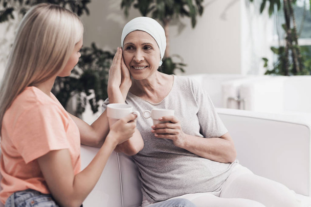 A woman with cancer is sitting on a white sofa next to her daughter. They sit with cups of tea in their hands. - Foto, Bild