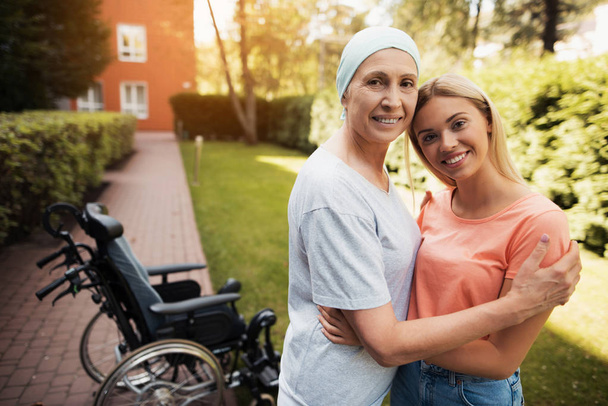 A woman with cancer stands up with her daughter. They embrace. Nearby is a wheelchair woman. - Valokuva, kuva