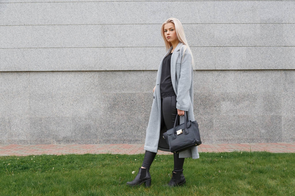 Модная женщина позирует на улице, держа в руках черную кожаную сумку, в стильных сапогах, элегантном пальто. Женская мода
. - Фото, изображение
