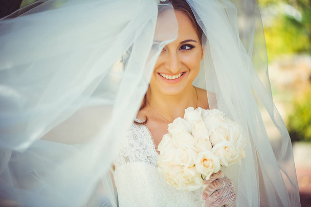 zarte glückliche Braut im Schleier, glückliche Frau im Brautkleid mit Blumenstrauß in den Händen, weißer Schleier bedeckt Gesicht, sanft und warm - Foto, Bild