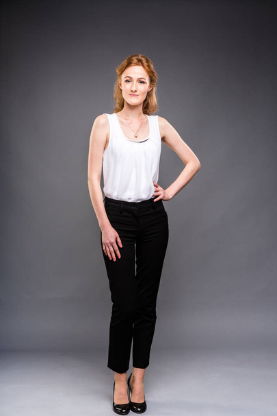 beautiful young Caucasian woman with long red hair in high heels, black trousers and a white shirt stands in full growth on a gray isolated background. - Fotografie, Obrázek