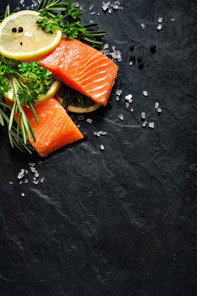 Fresh fish portions with lemon slices, rosemary, parsley, salt and peppercorns on a black stone background - Фото, зображення