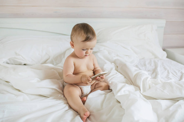 Little baby in a pampers plays with a smartphone on a bed - Foto, Imagem