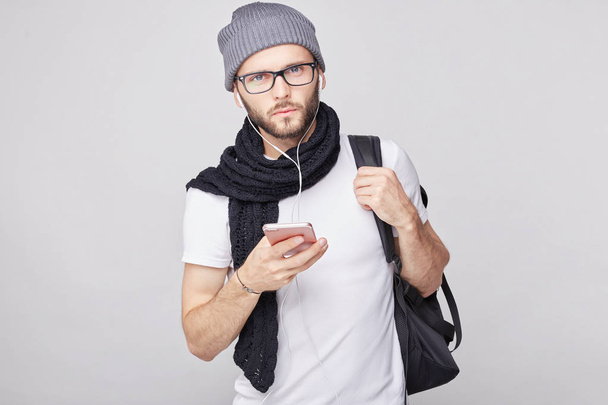 Stylish young bearded male blogger with backpack posing indoors while recording video on front-facing camera of his gadget, addressing himself to his followers and telling them about his road trip. - Photo, Image