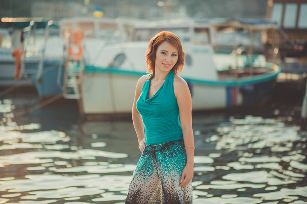 Amazing beautifull lady girl woman with red flame hair wearing fancy fashion green clothes posing sitting on wooden pier jetty wth port boats on background Adoring scene of sea mermaid - 写真・画像