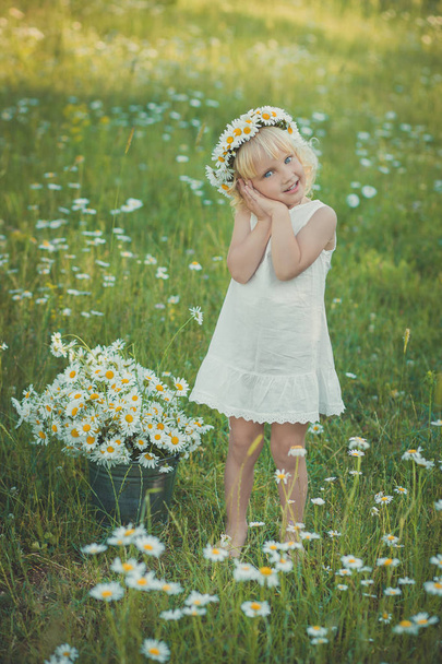 Cute young blond girl in white airy dress posing on daisy chamomile meadow with bouquet of flowers in hands basket metal bucket and wreath anademon top head smiling. - Photo, Image