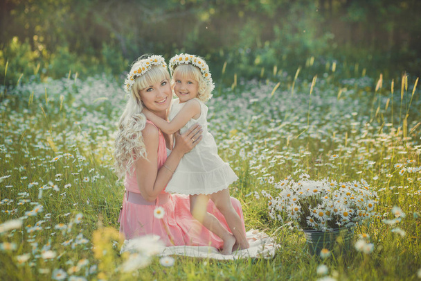 Magnificent picture of blond adorable lady woman mother mom in pink airy dress with daughter girl baby in white ambrace each other on daisy chamomile meadow with horse on background. - Valokuva, kuva