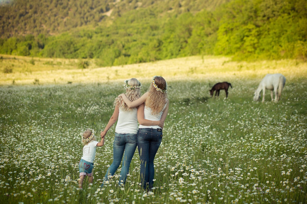 Twee jonge blonde dame in jeans en witte onderjas shirt lopen over weide van daisy kamille met meisje hand in hand dragen krans anadem van bloemen met paard en berg op achtergrond. - Foto, afbeelding