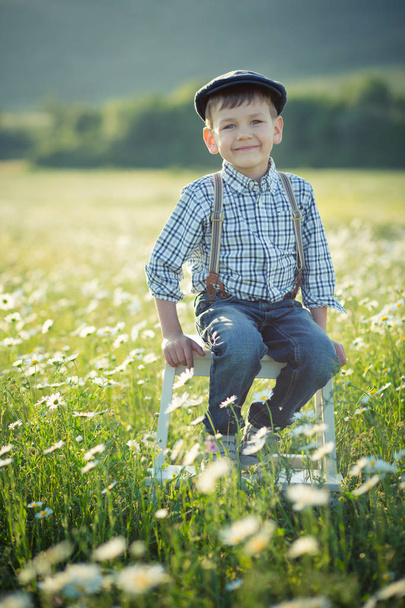 Cute handsome boy wearing jeans and shirt sitting on wooden chair on meadow of chamomiles daisy and smiling enjoying summer day in vallage farm. - Fotó, kép