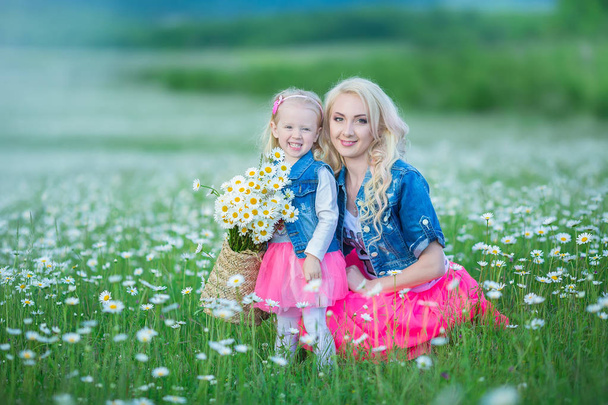 Mom and daughter on a picnic in the chamomile field. Two beautiful blondes wearing jeans jacket and pink skirt Mother and baby girl embracing in the chamomile field with basket of bouquet dandalion - Photo, Image
