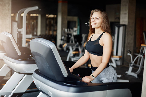 Portrait of crazy fit woman on a threadmill smiling looking at camera. Athletic coach warming up early in the morning in empty gym. - Zdjęcie, obraz