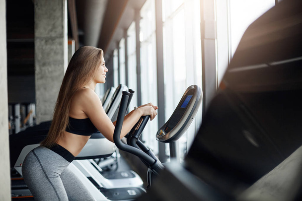 Portrait of beautiful female gym attendee running on elliptical cross trainer early in the morning. Workout before everyday job. - Foto, imagen