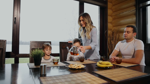Family of four having Breakfast in his kitchen with large Windows. People are smiling, mother kissing and hugging children. Mother father and two children. - Filmagem, Vídeo