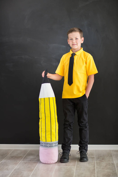 Cute handsome school boy in yellow t-shirt tie and stylish boots casuals standing cloase to black board with numbers and holding huge big soft toy pencil and smiling - Zdjęcie, obraz