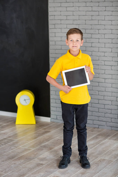 Chłopak ładny przystojny szkoły w żółty t-shirt tie i stylowe buty modne stały cloase na czarny deska z numerami i trzymając tablet i uśmiechając się - Zdjęcie, obraz