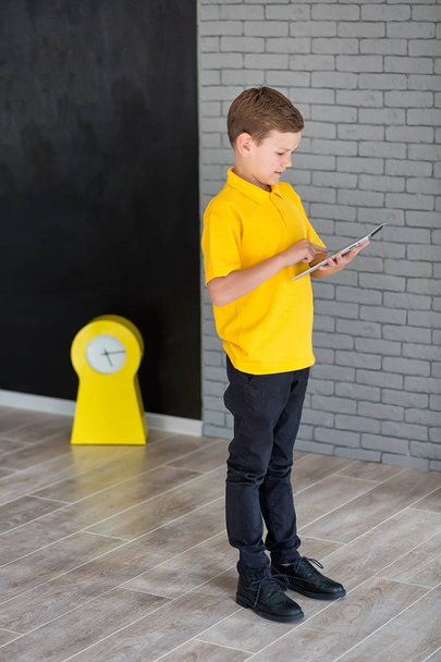 Cute handsome school boy in yellow t-shirt tie and stylish boots casuals standing cloase to black board with numbers and holding tablet and smiling - Фото, изображение