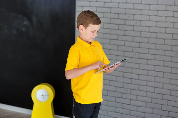 Cute handsome school boy in yellow t-shirt tie and stylish boots casuals standing cloase to black board with numbers and holding tablet and smiling - Photo, Image