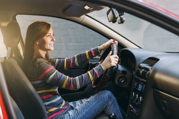 Beautiful young happy smiling European brown-haired woman with healthy clean skin dressed in a striped t-shirt sits in her red car with black interior. Traveling and driving concept. - Photo, Image