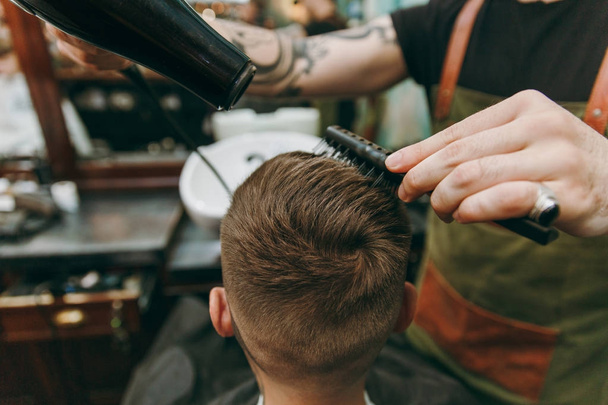 Tiro de perto de um homem a cortar o cabelo na barbearia. Cabeleireiro masculino em tatuagens que servem o cliente, secando o cabelo com um secador de cabelo
 - Foto, Imagem