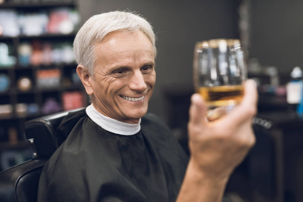 The old man drinks alcohol in the barber's chair in barbershop. - Foto, immagini