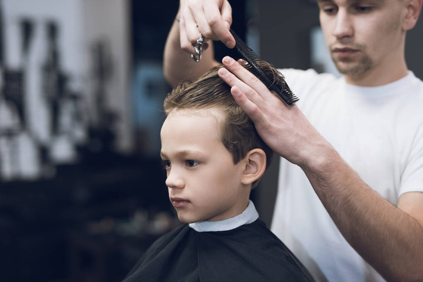 The hairdresser makes a fashionable pretty hairstyle for the boy in a modern barbershop. - Foto, Imagen