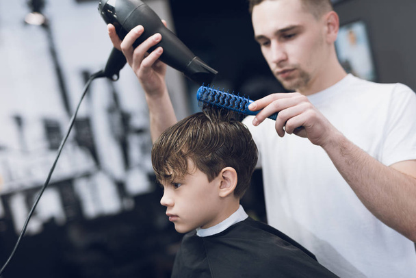 The hairdresser makes a fashionable pretty hairstyle for the boy in a modern barbershop. - Фото, изображение