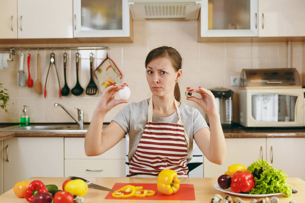 Una giovane donna attraente pensierosa in un grembiule sceglie tra uova di pollo e di quaglia in cucina. Il concetto di dieta. Uno stile di vita sano. Cucinare a casa. Preparare il cibo
. - Foto, immagini