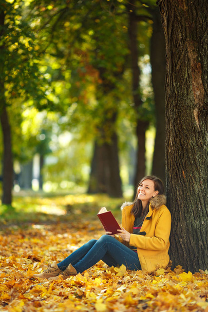 A beautiful happy smiling brown-haired woman in yellow coat and jeans sitting under the maple tree with a red book in fall city park on a warm day. Autumn golden leaves. Reading concept - Fotó, kép