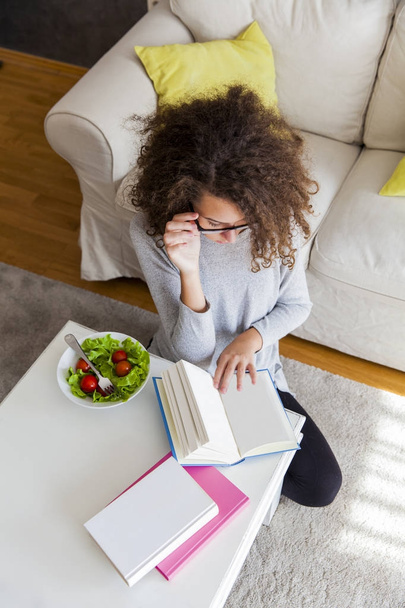 Curly hair teenage girl reading book and eating salad at home - 写真・画像