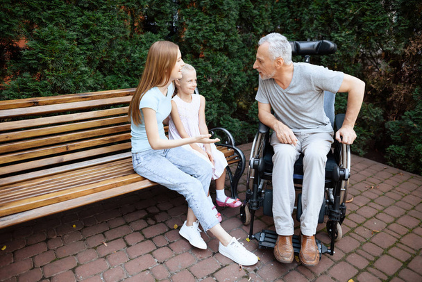 An elderly man is sitting in a wheelchair. He is seen by a woman with a girl. They are sitting on a bench in the park. - Photo, Image