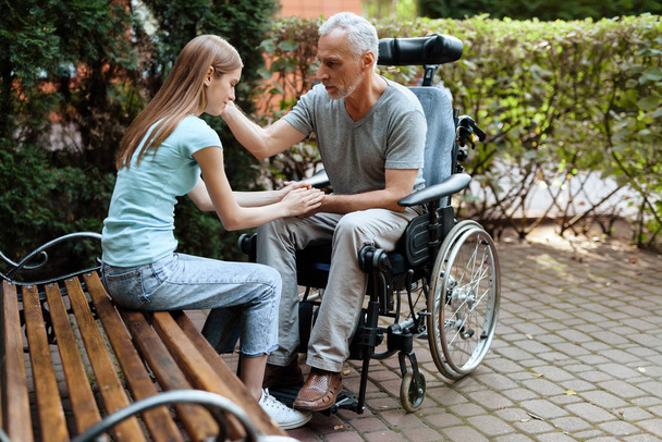 An elderly man is sitting in a wheelchair. Next to the bench is his daughter. They are holding hands. - Foto, Imagen