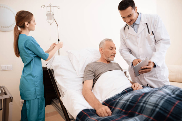 The old man lies on a cot in medical ward, and next to him stands a doctor. He is showing something to old man on tablet - Foto, Bild