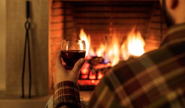 Man holding a glass of red wine on burning fireplace background - Foto, afbeelding
