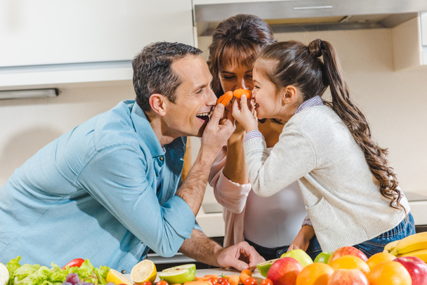 família feliz ao lado da mesa com frutas e legumes aplica pequenas cenouras ao nariz e olhando uns para os outros na cozinha
 - Foto, Imagem