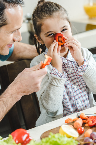 feliz padre con hija, niño pequeño aplica rodaja de pimienta a la nariz y mirando a la cámara en la cocina
 - Foto, Imagen