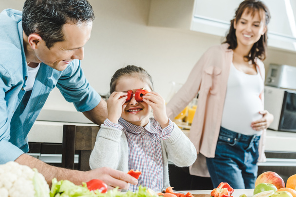 happy family, little kid applies pepper slices to eyes when father and pregnant mother watching on her at kitchen - Фото, зображення